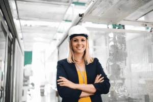 Woman in hard hat and business attire smiling confidently in a manufacturing environment, representing career opportunities in temporary roles within the manufacturing sector.