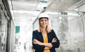 Smiling woman wearing a hard hat and professional attire, standing confidently in a manufacturing environment, representing career opportunities in the manufacturing sector.